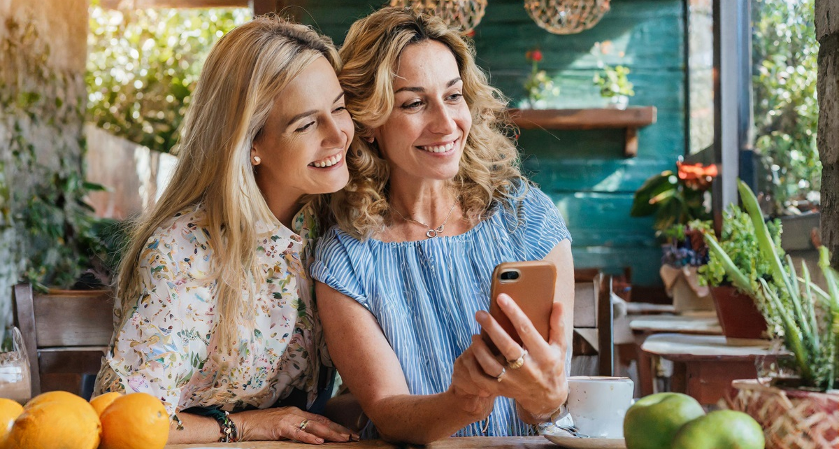 App-tly Ever After: My Daughter's Plan to Swipe Me into Romance 1348 tinywow Firefly mother with a cellphone with her adult daughter next to hear looking at the phone at ktichen 53221002