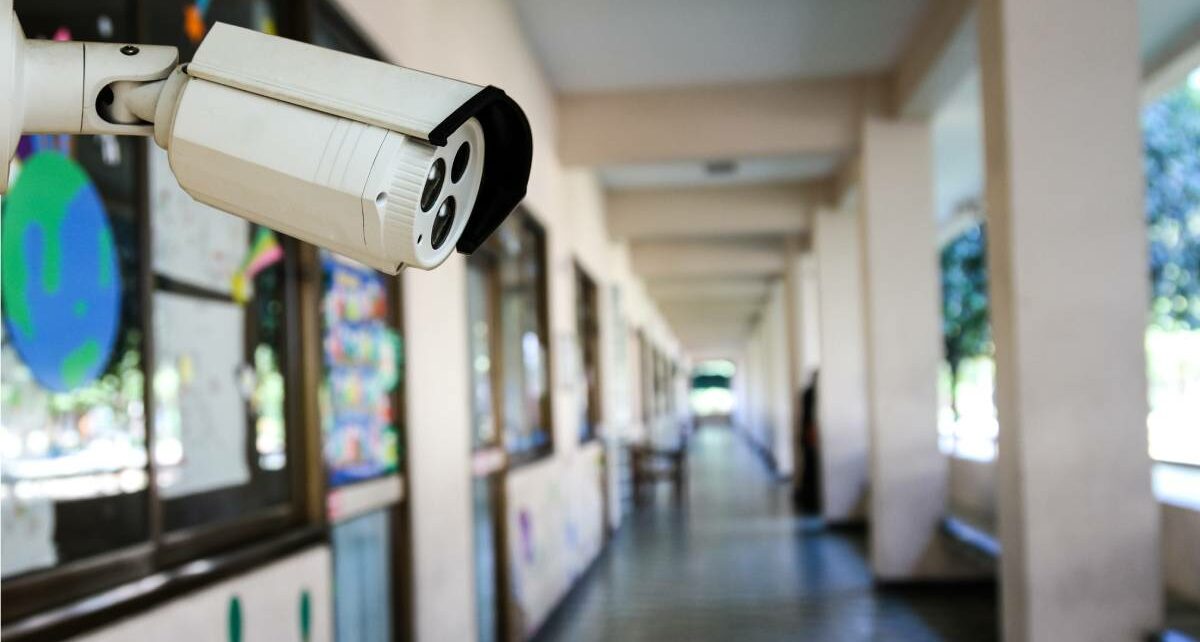 Close-up of a CCTV camera in a school hallway. The walls have posters and art. The other side of the hallway has benches.
