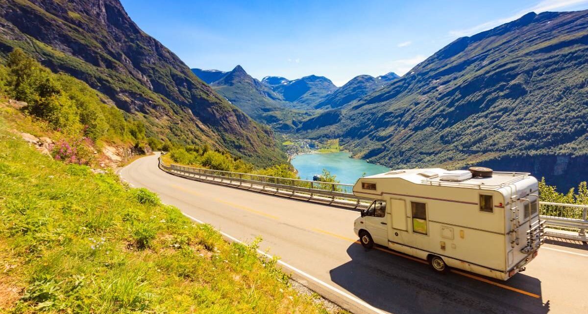 An RV traveling across a two-lane mountainous highway. There's a small lake, a valley, and the sun is shining.