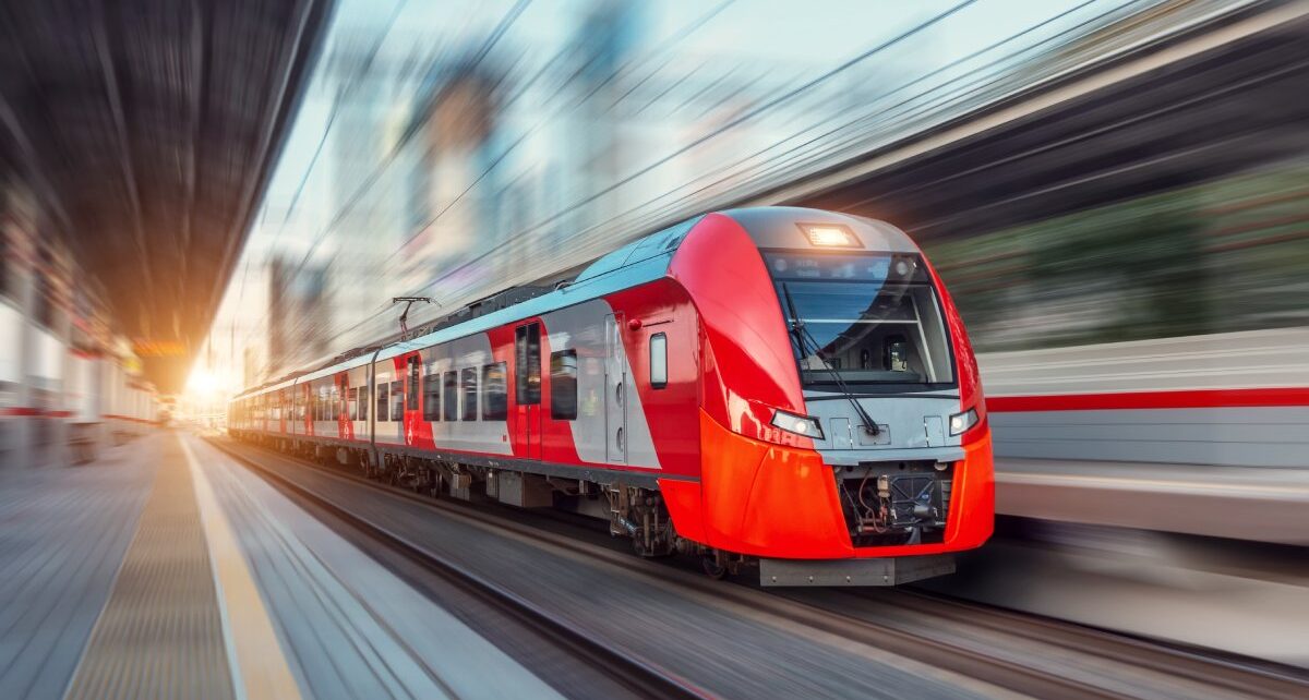 Why US Public Transportation Is So Expensive To Build 4 A red passenger train moves across the train tracks at a high speed. The train platform is empty.