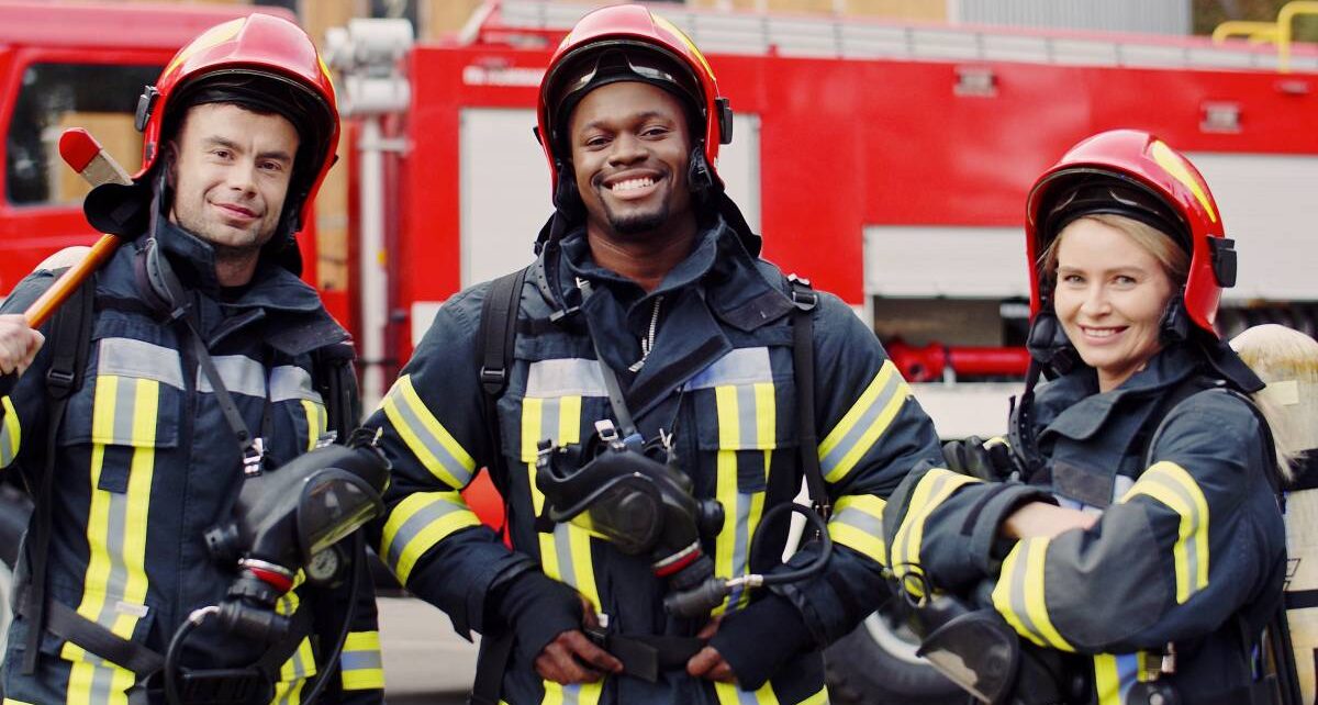 5 Pieces of Equipment Firefighters Use Every Day 114 Three firefighters dressed in black protective clothing and red helmets stand in front of a fire truck and smile.