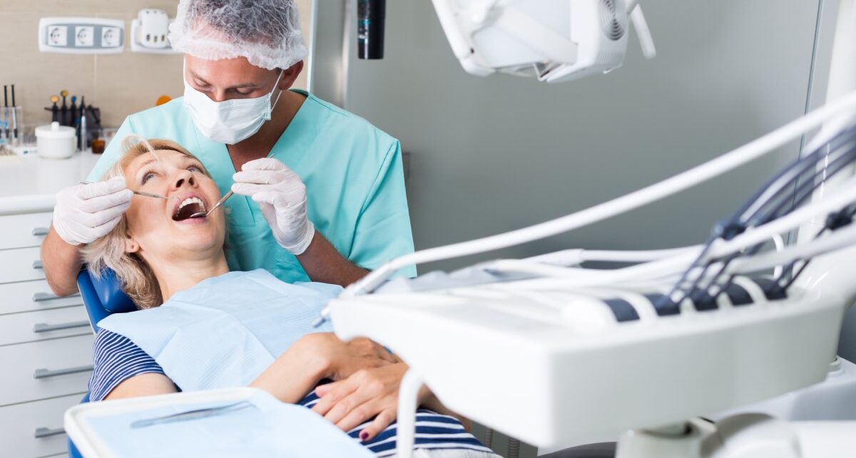 A male dentist wearing gloves and a face mask using stainless steel tools to treat a female patient's mouth.