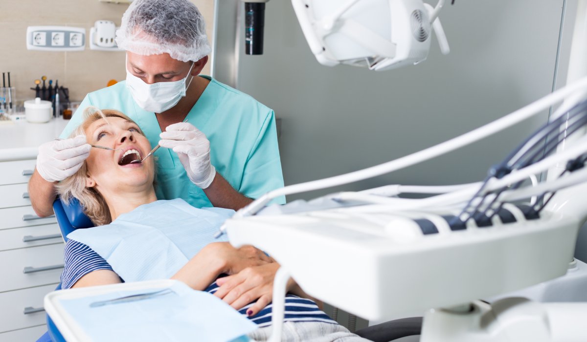 A male dentist wearing gloves and a face mask using stainless steel tools to treat a female patient's mouth.