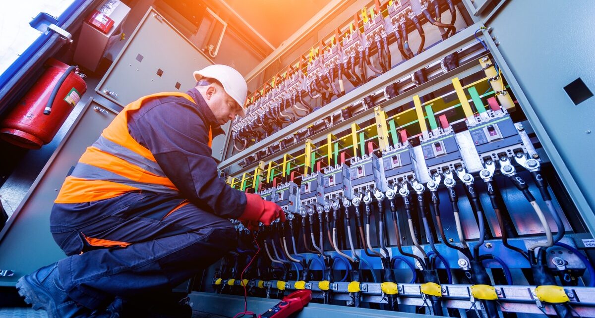 What Every Electrical Maintenance Program Needs 22 A ground-view of an electrician in a hard hat and orange high-visibility vest inspecting an electrical cabinet.
