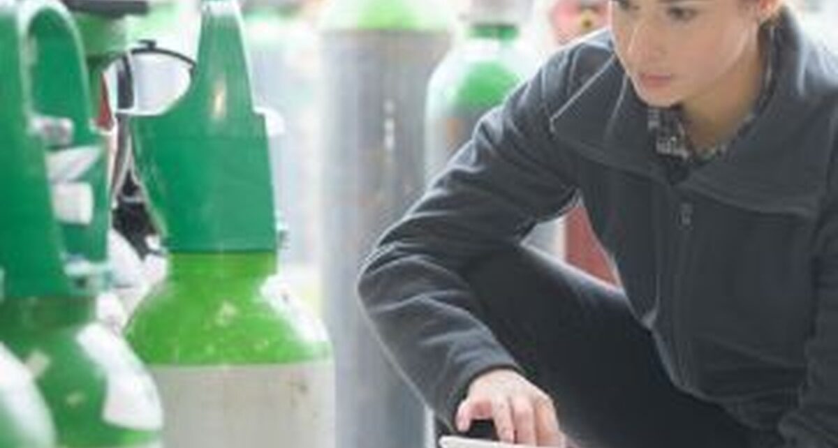 Best Practices for Long-Term Medical Gas Storage 1 Person in a dark jacket crouching with a tablet while inspecting several large green and white gas cylinders indoors.