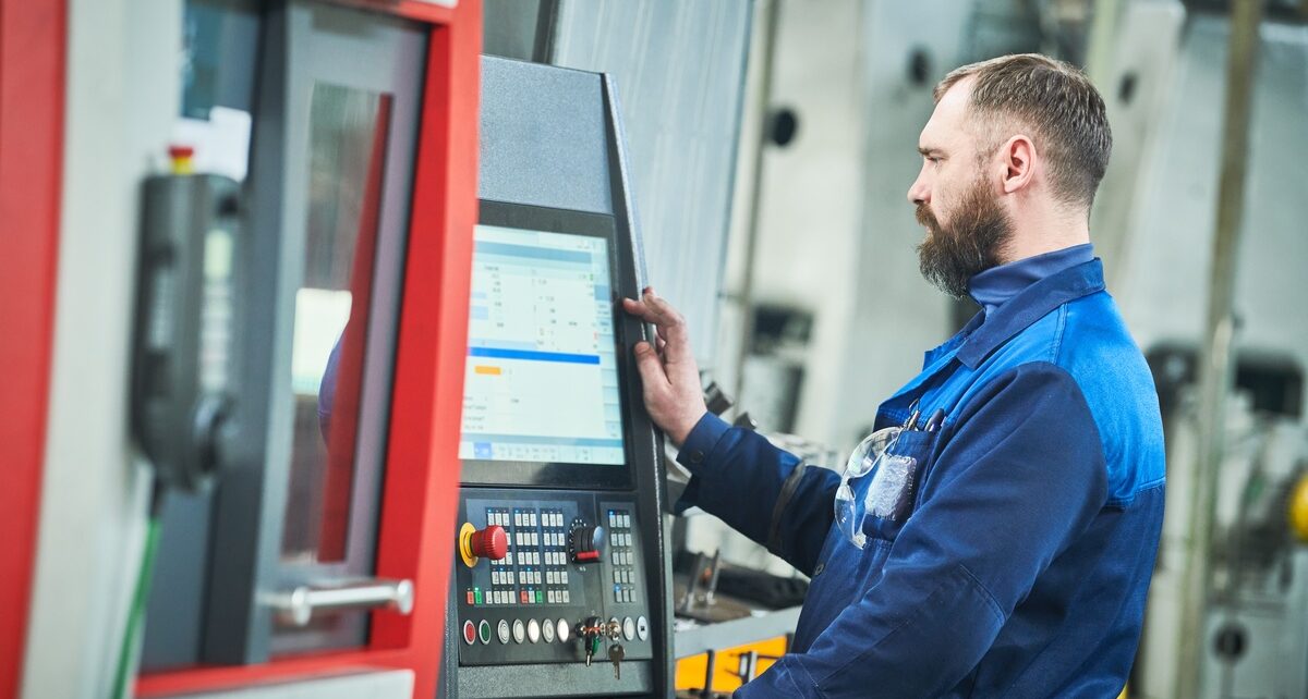 How 5-Axis CNC Machining Is Revolutionizing Industries 75 A CNC machine operator stands at the computer in full uniform. The machine is to their left and the uniform is blue.