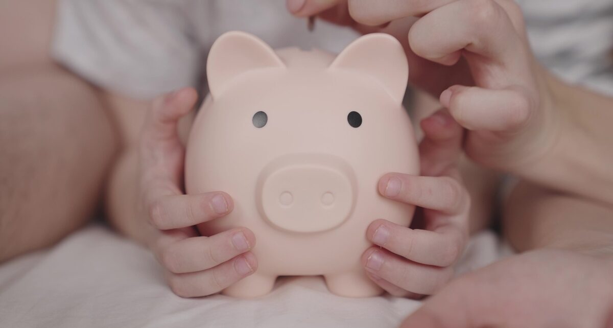 A child holding a light pink piggy bank between their hands while an adult drops a coin into the coin slot.