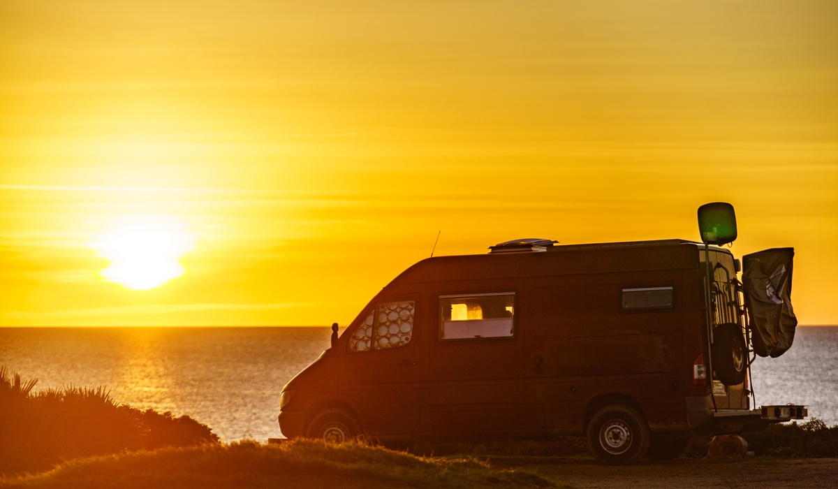 A wide view of a camper van parked at the edge of a cliff, overlooking the ocean as the sun sets on the horizon.