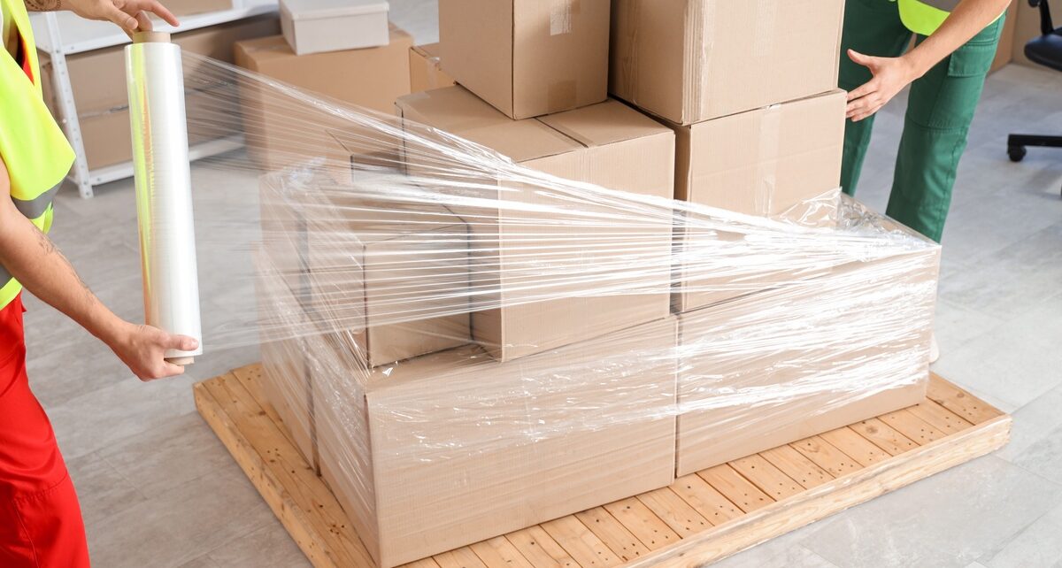 The Pros and Cons of Manual Stretch Wrapping 1 A wooden pallet with cardboard boxes stacked on it being manually wrapped with stretch wrap by two workers.