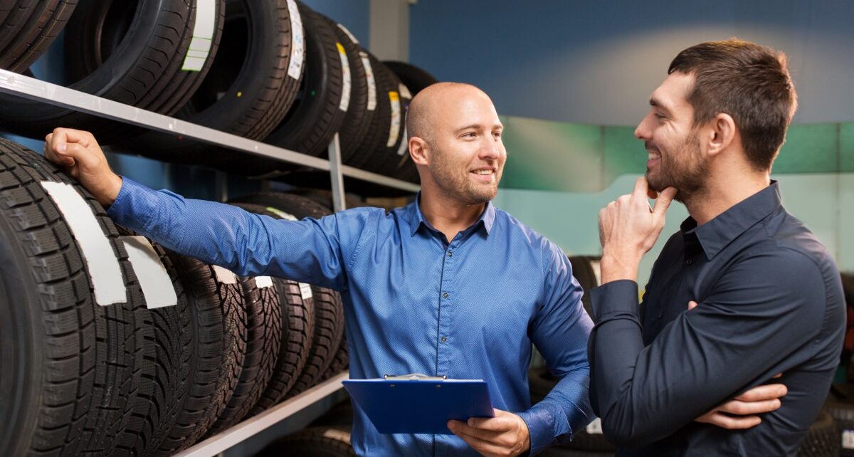 What’s Most Important When Shopping for New Car Tires 3 A salesman in a blue shirt with a clipboard touches a rack of new car tires while talking to a male customer.