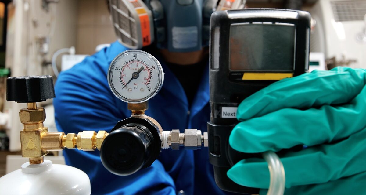 Choosing the Best Calibration Gas Canister Size 1 A close-up view shows a person wearing a gas mask and teal gloves using a device to calibrate a gas canister.