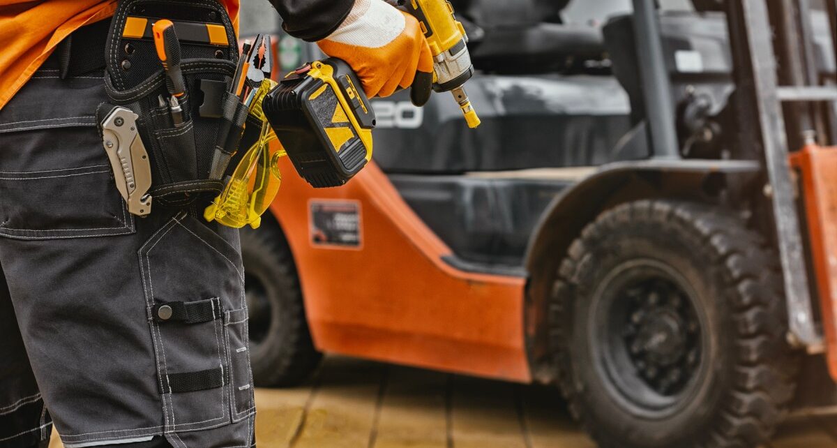 Troubleshooting Common Problems with Heavy Equipment 4 A worker in an industrial facility standing in front of an orange forklift. The worker has a drill in their right hand.