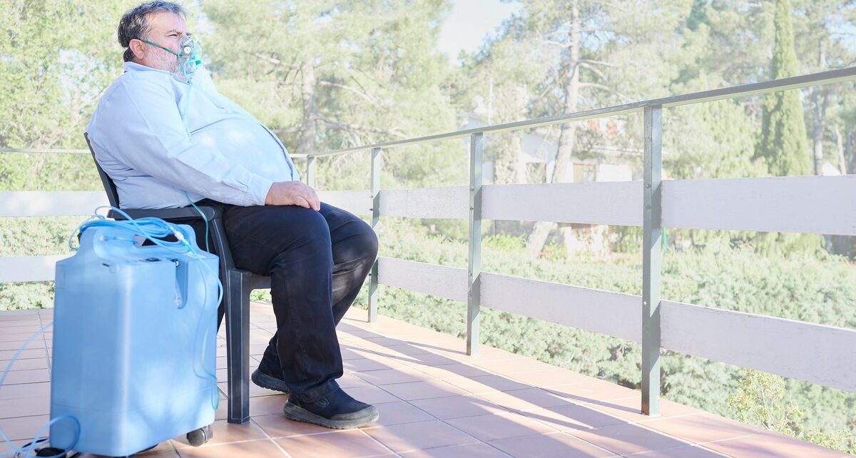Why Oxygen Concentrators Are Beneficial for Seniors' Health 99 A man sitting on a chair outdoors wearing an oxygen mask with a concentrator on his side, looking at a forest.