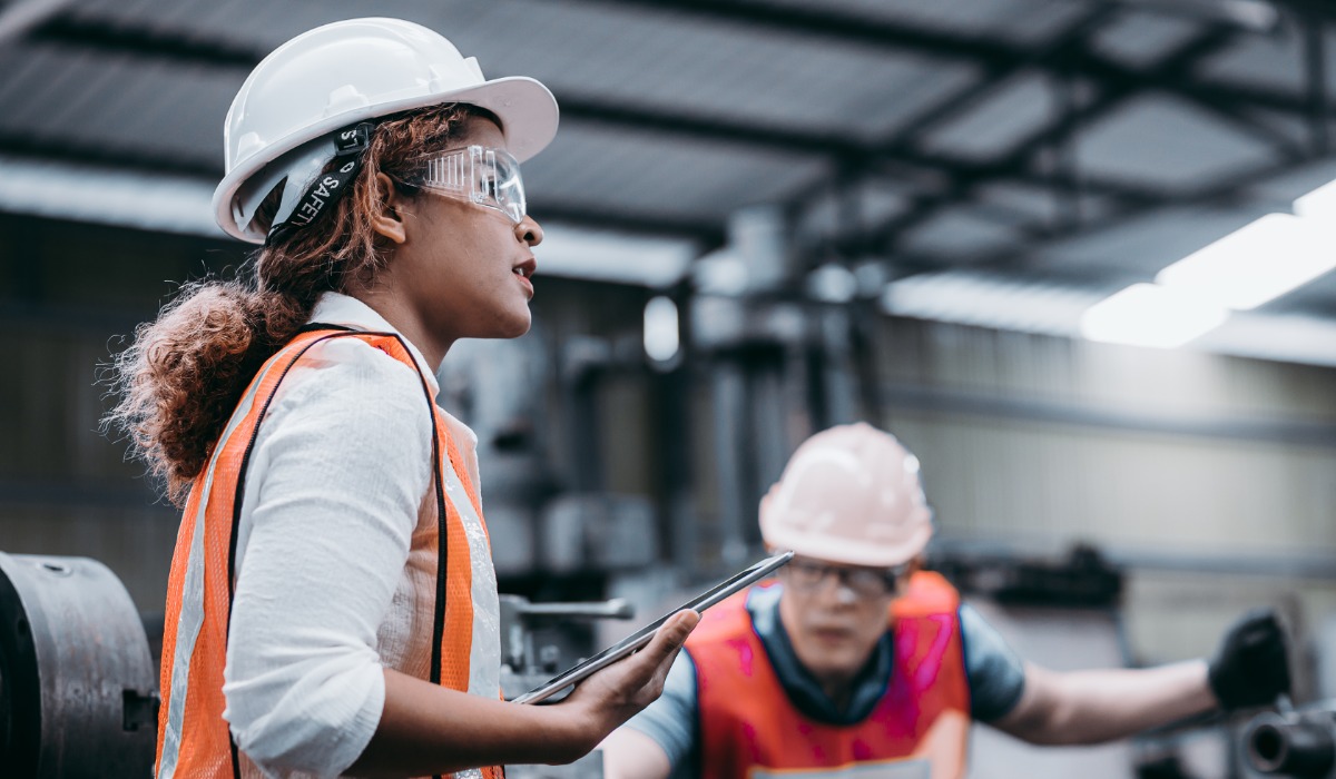 Two engineers wearing safety goggles and hard hats in an industrial work environment. One engineer holds a tablet.