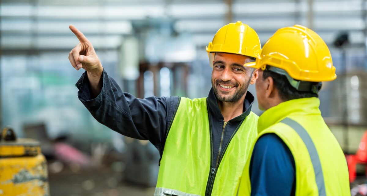 Signs You Need to Replace Your Finishing or Tumbling Media 2 Engineers wearing hard hats and safety vests. One engineer points at a machine while smiling at the other worker.