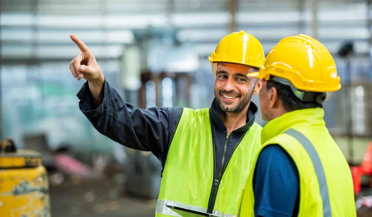 Engineers wearing hard hats and safety vests. One engineer points at a machine while smiling at the other worker.
