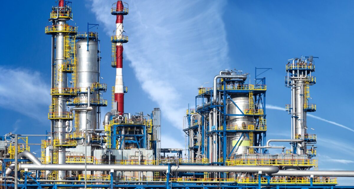 Aerial view of an oil refinery with silver distillation towers, pipelines, and yellow scaffolding against a bright blue sky.
