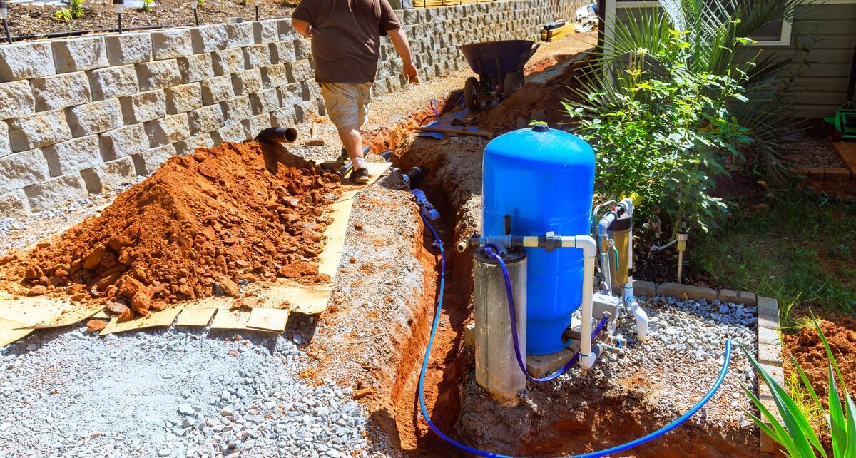 What You Need To Install a Well on Your Property 113 A man in a brown shirt in a resiential backyard walking away from a well pump system near a tench and mount of dirt.