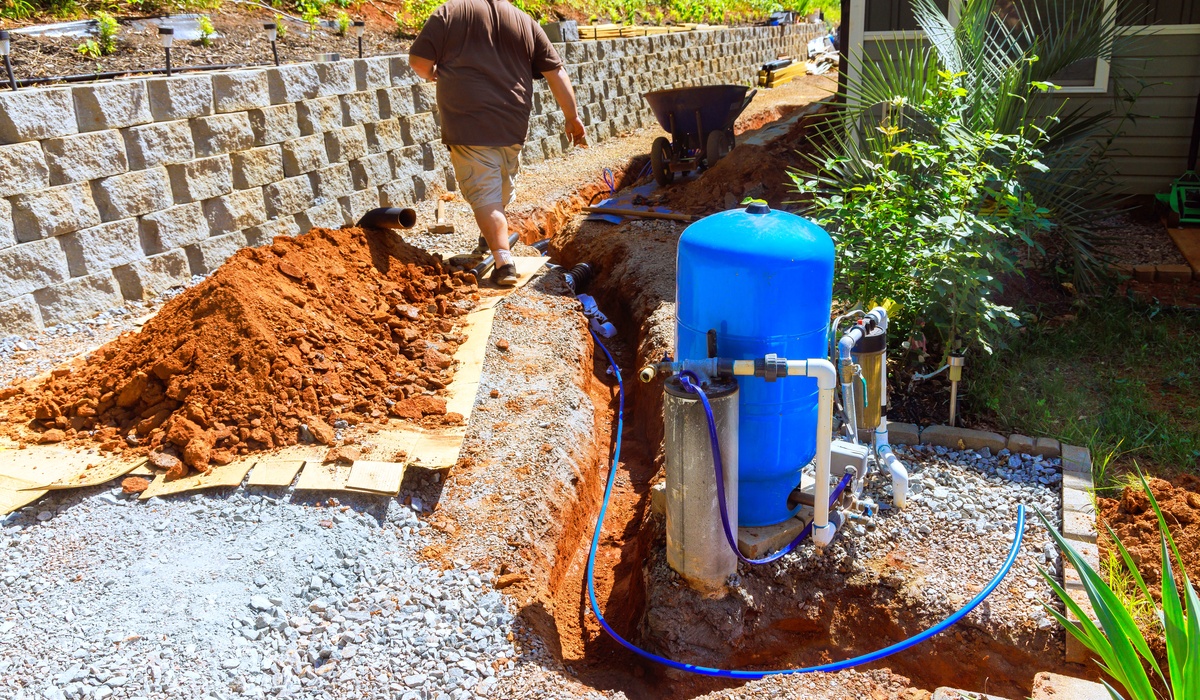 A man in a brown shirt in a resiential backyard walking away from a well pump system near a tench and mount of dirt.