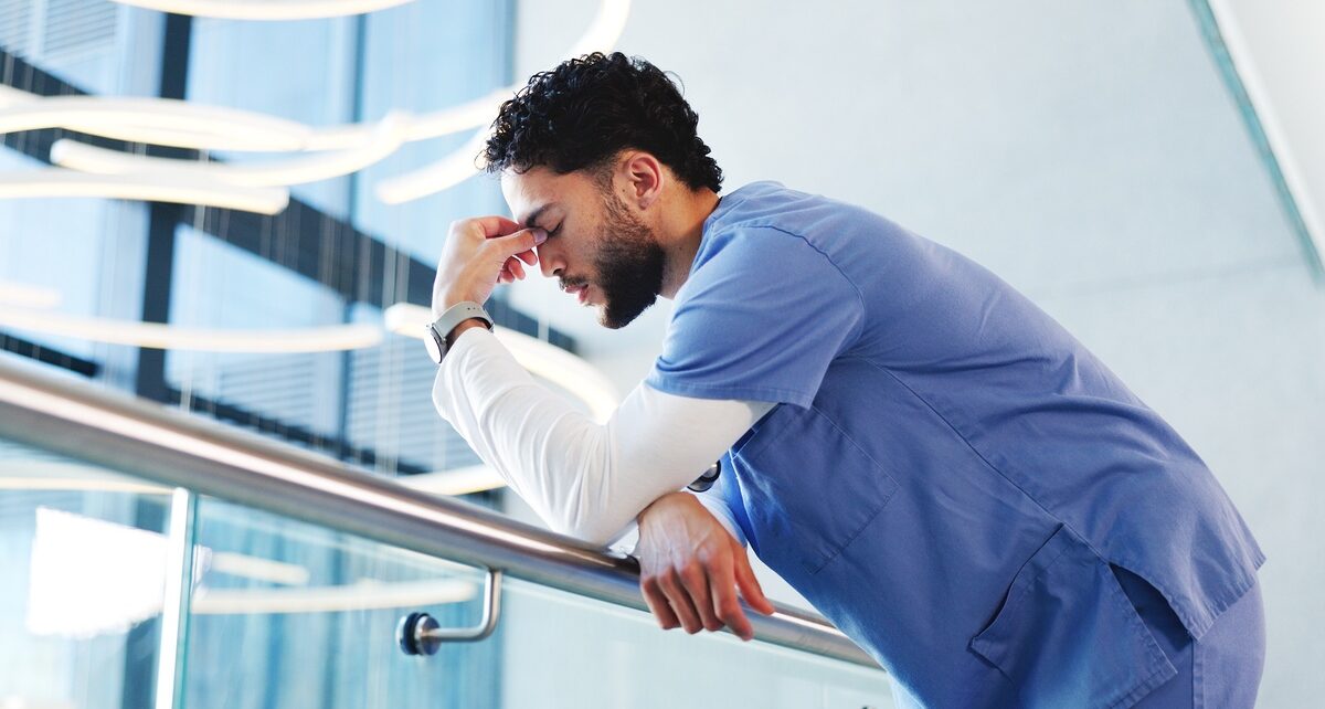 Burnout at the Bedside: The Hidden Patient Safety Crisis 94 A male nurse in blue scrubs leans against a hospital railing, head bowed and hand pressed to his forehead in exhaustion.