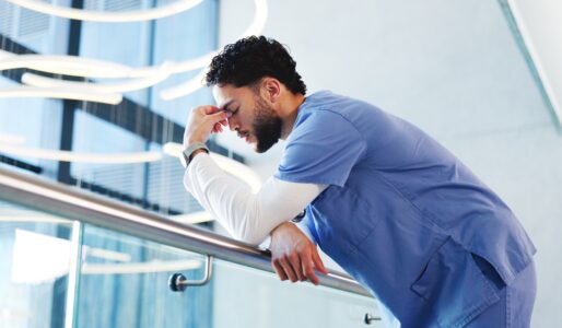 A male nurse in blue scrubs leans against a hospital railing, head bowed and hand pressed to his forehead in exhaustion.