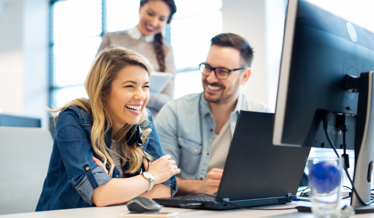 A man and woman are laughing while they sit at a desk looking at an office computer. A smiling woman stands behind them.