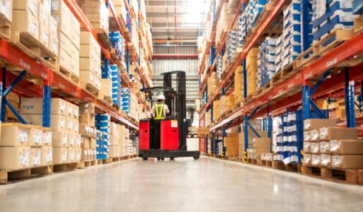 A worker driving a red forklift down a warehouse aisle between tall racks stacked with boxes and pallets.